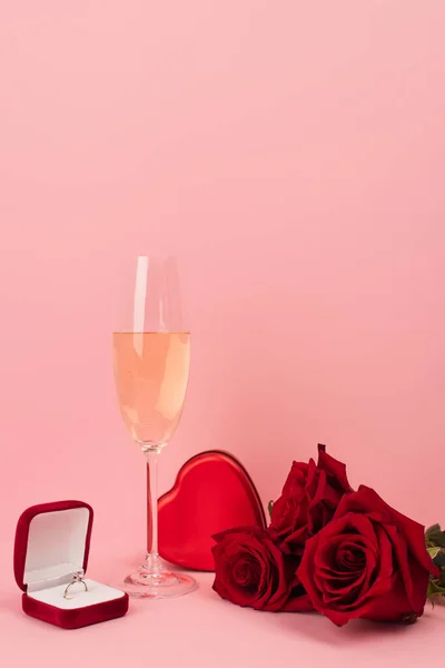 Copo de champanhe perto de caixa em forma de coração, rosas e anel de noivado em rosa — Fotografia de Stock