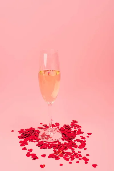 Glass of champagne near red heart-shaped confetti on pink — Stock Photo