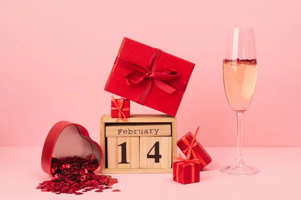 Coffrets cadeaux rouges près du calendrier en bois avec lettrage 14 février près du champagne en verre et confettis sur rose — Photo de stock