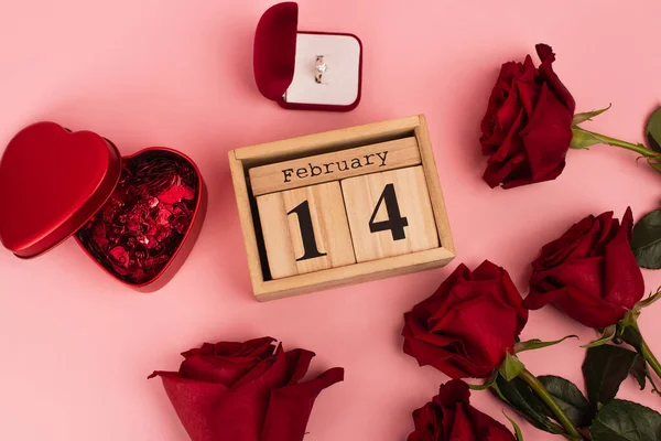 Top view of red roses near calendar with 14 february lettering, confetti and engagement ring on pink — Stock Photo