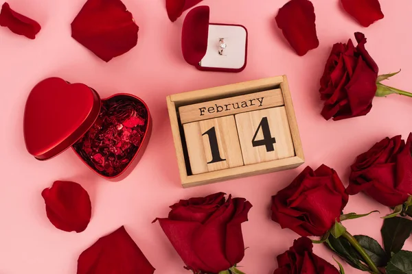 Vue du haut des roses rouges près du calendrier avec 14 février, confettis et bague de fiançailles autour des pétales sur rose — Photo de stock
