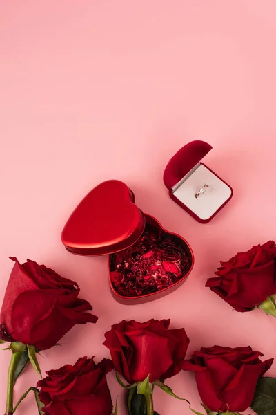 Top view of red roses near box with heart-shaped confetti and engagement ring on pink — Stock Photo