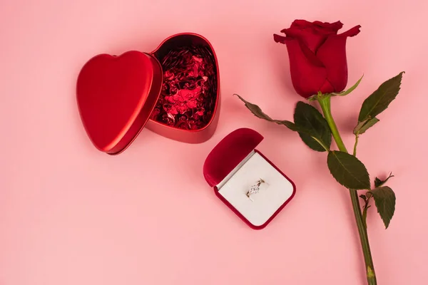 Вид зверху на червону троянду біля коробки з серцеподібною конфетті і кільцем заручин на рожевому — стокове фото