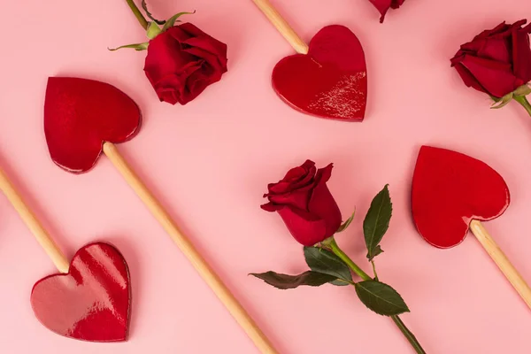 Вид зверху на серцеподібні льодяники біля червоних троянд на рожевому — стокове фото