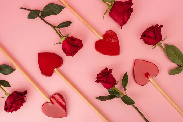 Disposición plana de piruletas en forma de corazón cerca de rosas rojas en rosa - foto de stock