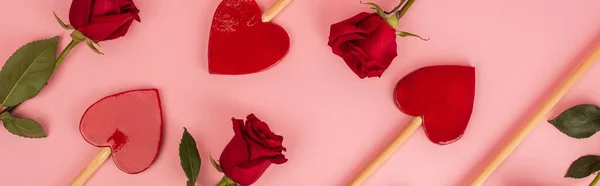 Disposición plana de piruletas en forma de corazón cerca de rosas rojas en la bandera rosa - foto de stock