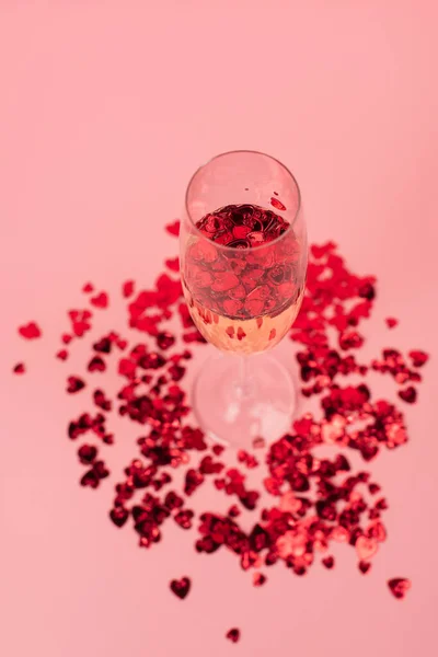 High angle view of glass with champagne and red heart-shaped confetti on pink — Stock Photo