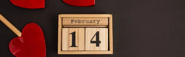 Top view of wooden calendar with 14 february lettering near heart-shaped lollipops on black, banner — Fotografia de Stock