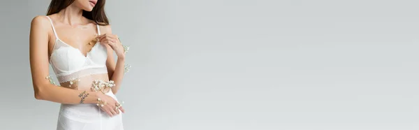 Vue partielle de femme séduisante avec des fleurs blanches de gypsophile sur le corps posant en soutien-gorge isolé sur gris, bannière — Photo de stock
