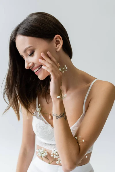 Morena en sujetador blanco, con pequeñas flores en el cuerpo, sonriendo aislada en gris - foto de stock