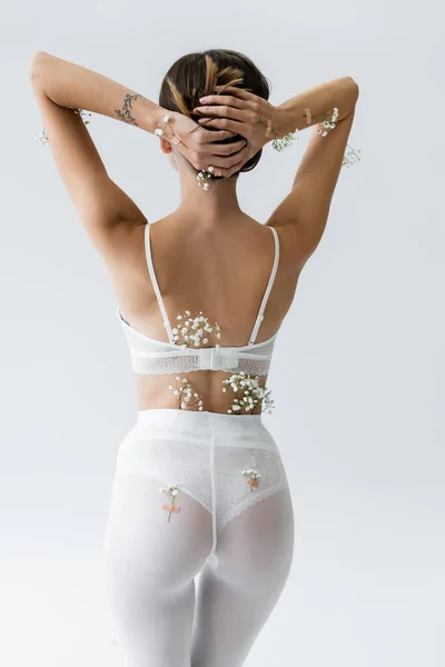 Back view of slim woman in white bra and tights, with tiny flowers on body, posing with hands behind head isolated on grey — Stock Photo