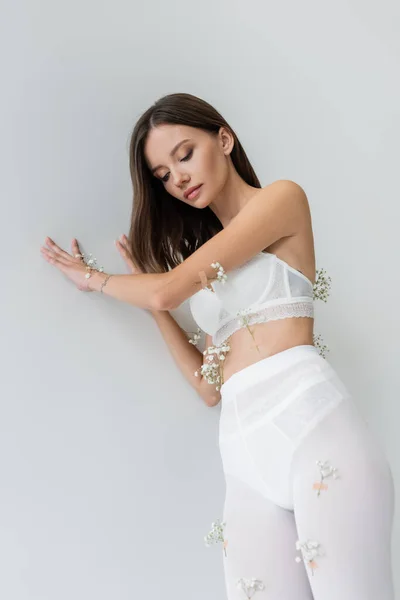 Low angle view of sexy woman in white tights and bra, decorated with gypsophila flowers, posing on grey background — Stock Photo