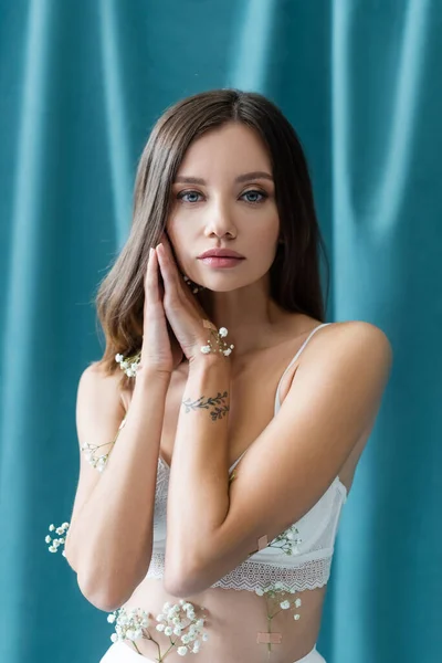 Jovem mulher tatuada em sutiã branco, com pequenas flores no corpo, posando com as mãos perto do rosto perto de cortinas verdes no fundo — Fotografia de Stock