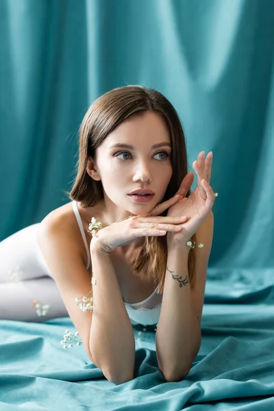 Sensual tattooed woman with white tiny flowers on body looking away while lying on green cloth — Stock Photo