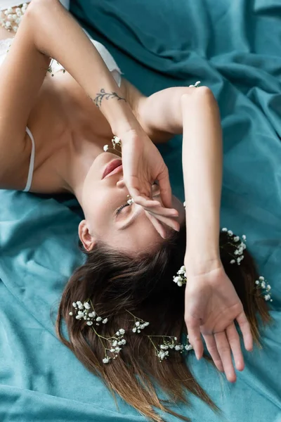 Vista aerea della donna con fiori di gypsophila bianchi sul corpo e sui capelli, sdraiata con gli occhi chiusi sul drappeggio turchese — Foto stock