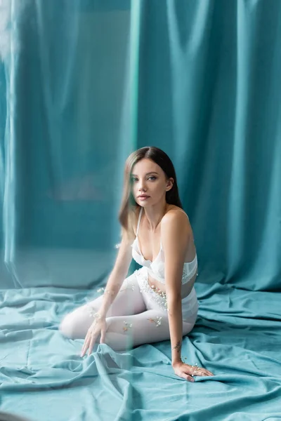 Junge Frau in Spitzen-BH und weißen Strumpfhosen, mit kleinen Blüten am Körper, die in die Kamera schaut, während sie auf grünem Vorhang sitzt — Stockfoto