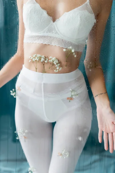 Vista ritagliata di donna sexy in collant bianchi e reggiseno di pizzo, con fiori di gypsophila sul corpo, posa vicino vetro bagnato su sfondo verde — Foto stock
