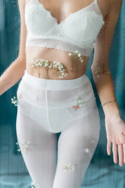 Vue recadrée de femme sexy en collants blancs et soutien-gorge en dentelle, avec des fleurs de gypsophile sur le corps, posant près de verre mouillé sur fond vert — Photo de stock