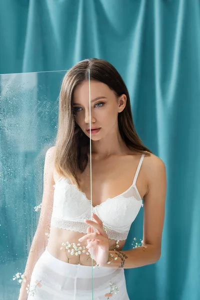 Young brunette woman in lace bra, with tiny gypsophila flowers on body, standing near wet glass and turquoise drapery on background — Stock Photo
