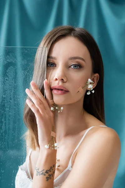 Jeune femme séduisante avec des fleurs blanches minuscules sur le corps en regardant la caméra près de verre humide sur fond drapé vert — Photo de stock