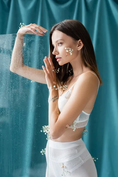 Sensual woman in white lace bra, with gypsophila flowers on body, looking away near wet glass on green draped background — Stock Photo