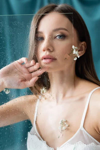 Jolie jeune femme, décorée avec des fleurs de gypsophila, regardant la caméra près de verre mouillé et draperie verte sur fond — Photo de stock