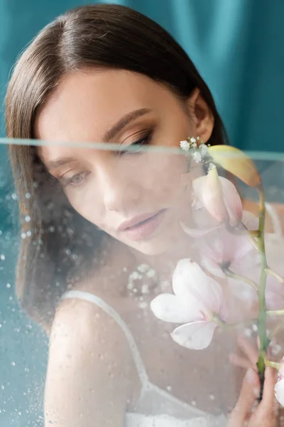 Mujer joven y sensual con orquídea blanca cerca de vidrio húmedo borroso sobre fondo turquesa - foto de stock