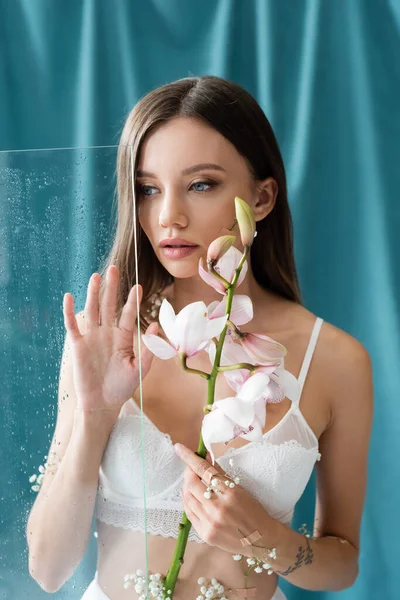 Sexy jovem mulher com branco orquídea tocando molhado vidro perto verde drapejar no fundo — Fotografia de Stock