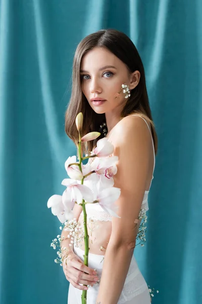 Brunette woman with white gypsophila flowers on body looking at camera while holding orchid near turquoise draped background — Stock Photo