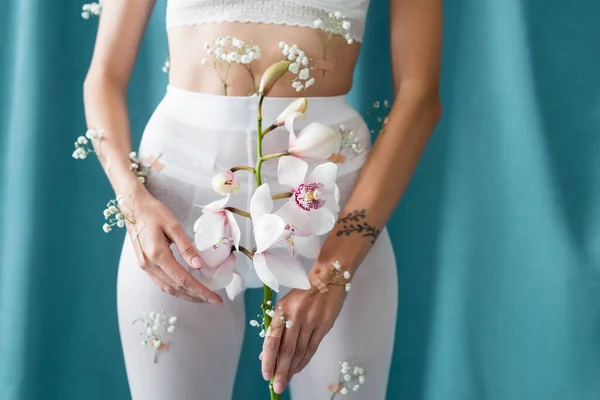 Vista recortada de mujer delgada en medias blancas con flores de gypsophila que sostienen la orquídea sobre fondo verde drapeado - foto de stock