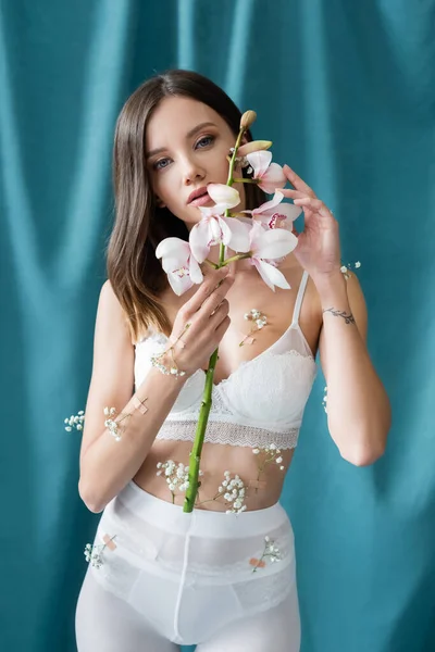 Charming woman with white orchid and body decorated with gypsophila flowers looking at camera on green draped background — Stock Photo