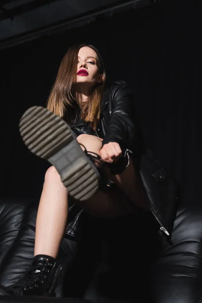 Низкий угол зрения сексуальной женщины регулируя ботинок, сидя на диване на черном фоне — стоковое фото