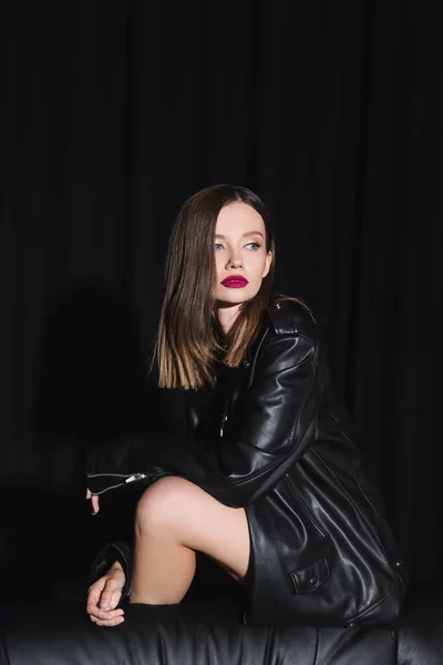 Sensual brunette woman in leather jacket looking away while posing on black background with drape — Stock Photo