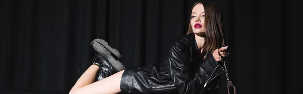 Sexy brunette woman in leather jacket and boots posing with handcuffs while lying on black draped background, banner — Stock Photo