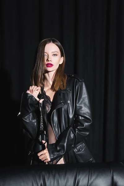 Hot woman in leather jacket and lace bodysuit looking at camera while holding handcuffs on black background with drapery — Stock Photo