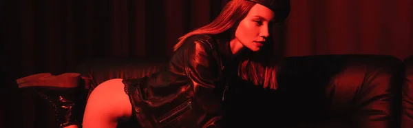 Sexy donna in giacca di pelle e stivali in posa sul divano a luce rossa su sfondo nero, banner — Foto stock