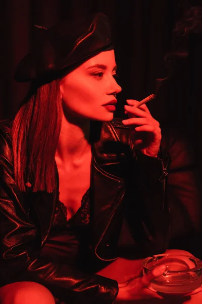 Sexy woman in leather beret, jacket and bodysuit smoking in red light on black background — Stock Photo