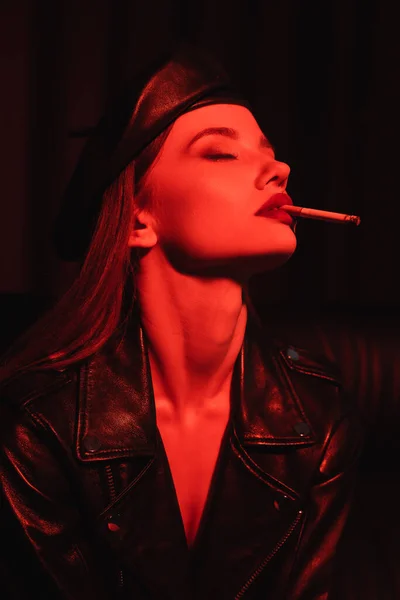 Seductive woman in leather jacket and beret smoking with closed eyes in red light on black background — Stock Photo