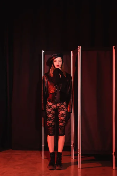 Visão de comprimento total da jovem mulher em leggings de renda, botas e jaqueta de couro em pé perto da tela de privacidade no fundo preto com cortinas — Fotografia de Stock