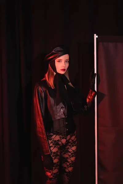 Glamour woman in leather jacket and lace leggings looking at camera near privacy screen on black background — Stock Photo