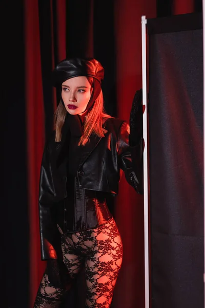 Sexy woman in leather jacket, lace leggings, beret and gloves looking away near privacy screen on dark draped background — Stock Photo
