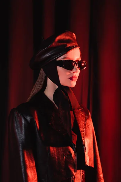 Стильная женщина в черных солнцезащитных очках, кожаной куртке и берете стоит на темном фоне с драпировкой и красным светом — стоковое фото