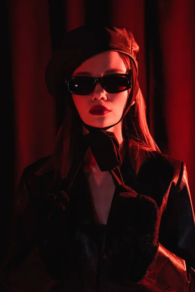 Glamour woman in leather beret, black jacket, sunglasses and gloves posing on dark draped background — Stock Photo