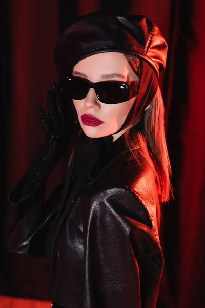 Stylish woman in leather jacket and beret adjusting black sunglasses on dark background with drape — Stock Photo