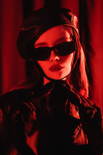 Portrait of young woman in black sunglasses, leather beret and gloves on dark background with red light — Stock Photo