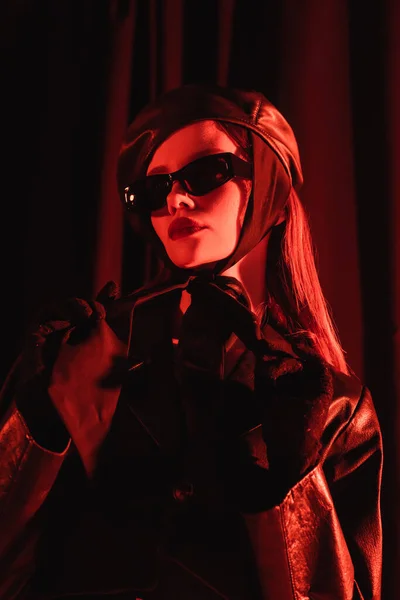 Low angle view of glamour woman in black sunglasses, leather beret and gloves standing on dark background with red light — Stock Photo