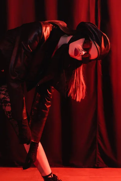 Jovem mulher de jaqueta de couro preto, luvas, boina e óculos escuros posando no fundo escuro com drapeados e luz vermelha — Fotografia de Stock