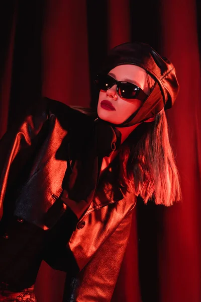 Mujer glamour en gafas de sol negras, chaqueta de cuero y boina posando sobre fondo oscuro con cortinas y luz roja - foto de stock