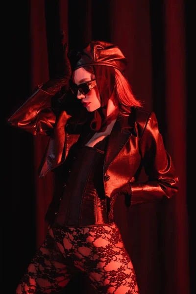 Mulher sexy em leggings de renda, jaqueta de couro preto e óculos de sol posando com a mão no quadril no fundo escuro drapeado — Fotografia de Stock