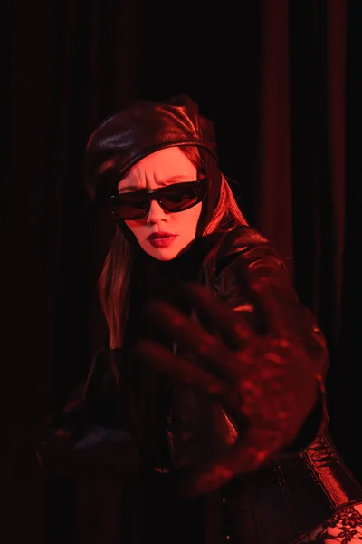 Frowning woman in black sunglasses, beret and leather jacket showing refuse gesture on dark background — Stock Photo